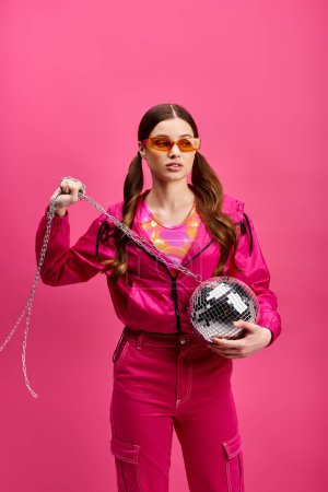 Une femme vibrante d'une vingtaine d'années, revêtue d'une élégante tenue rose, tient une boule disco, exsudant de l'énergie sur fond rose.