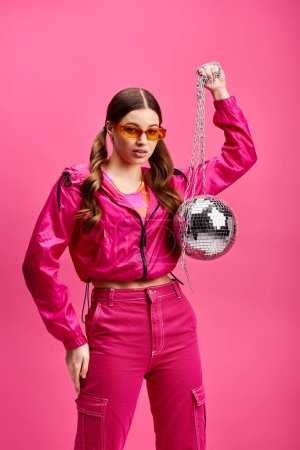 A stylish young woman in her 20s wears a pink outfit while holding a disco ball in a studio with a pink background.