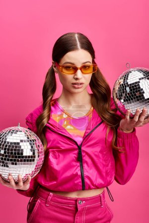 Stylish woman in her 20s, wearing a pink jacket, holds two shimmering disco balls in a vibrant studio setting.
