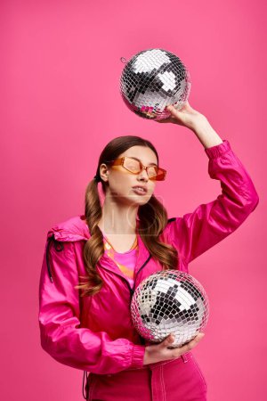 Foto de Young woman in her 20s, stylish in a pink jacket, holds two disco balls against a vibrant pink background. - Imagen libre de derechos