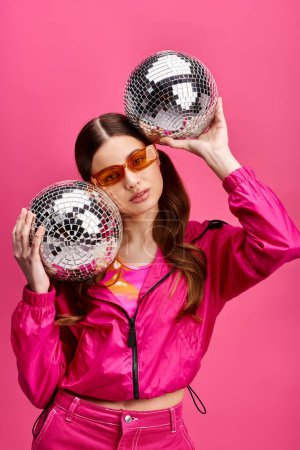 Téléchargez les photos : A stylish woman in her 20s, clad in a pink jacket, holds two disco balls in a studio with a vibrant pink background. - en image libre de droit