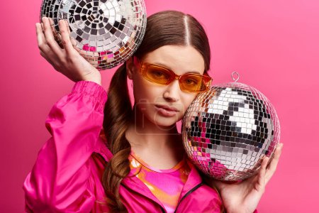 A young, stylish girl in her 20s wearing a pink jacket holds two disco balls in a studio with a pink background.