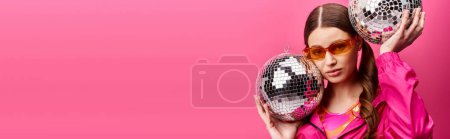 Téléchargez les photos : A young stylish woman in her 20s, wearing a pink shirt, joyfully holding two disco balls in a studio with a pink background. - en image libre de droit