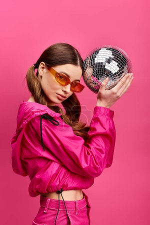 A stylish woman in her 20s wearing a pink jacket, holding a shimmering disco ball in a studio with a pink background.