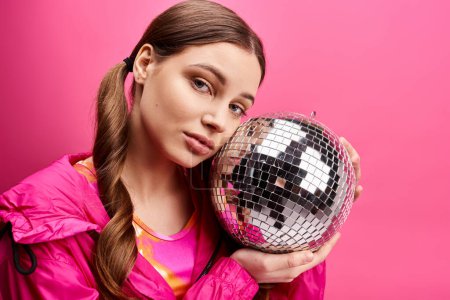 Téléchargez les photos : A young woman in her 20s holds a disco ball in front of her face, radiating a sparkling and glamorous aura against a pink background. - en image libre de droit