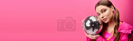 Junge Frau in den Zwanzigern hält Discokugel vor ihr Gesicht und schafft ein skurriles und geheimnisvolles Spektakel vor rosa Hintergrund.