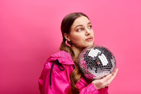 Téléchargez les photos : A stylish young woman in her 20s wearing a pink jacket, holding a sparkling disco ball in a studio with a pink background. - en image libre de droit