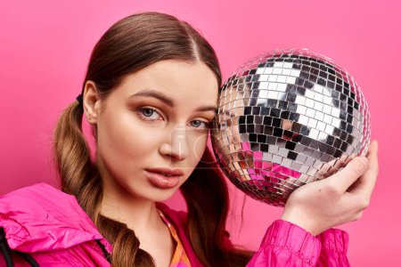 Photo for A stylish young woman in her 20s holds a disco ball in front of her face, creating a captivating reflection. Pink background in studio. - Royalty Free Image