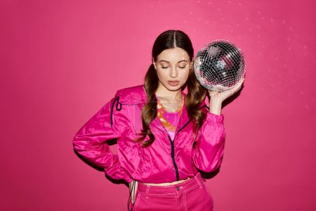 Una joven de unos 20 años, con estilo y chaqueta rosa, sostiene una bola de discoteca en un estudio con un vibrante fondo rosa.