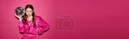 Téléchargez les photos : Une femme élégante d'une vingtaine d'années, vêtue d'une veste rose, pose gracieusement tout en tenant une boule disco sur un fond rose vif. - en image libre de droit