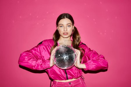 Téléchargez les photos : Stylish young woman in her 20s, wearing a pink jacket, holding a disco ball, against a vibrant pink background. - en image libre de droit