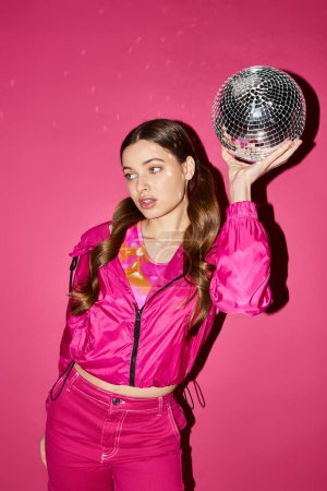 A stylish woman in her 20s, wearing a pink jacket, holding a disco ball in a studio with a pink background.