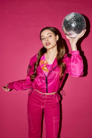 Une femme élégante dans la vingtaine tient une boule disco jusqu'à son visage dans un studio avec un fond rose, créant un reflet éblouissant.