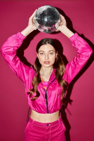 Photo for A stylish young woman in her 20s holds a disco ball above her head in a studio with a pink background. - Royalty Free Image