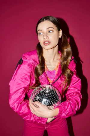 A stylish young woman in her 20s wearing a pink jacket holds a disco ball in a studio with a pink background.