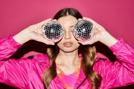 Téléchargez les photos : Young woman in her 20s, stylishly dressed in a pink jacket, holding two disco balls in a vibrant studio setting. - en image libre de droit