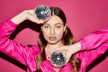 Una mujer joven y elegante de unos 20 años sosteniendo dos bolas de discoteca en un estudio con un fondo rosa.