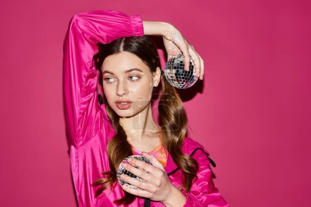 Foto de A stylish young woman in her 20s wearing a pink outfit holds a dazzling disco ball in a studio with a pink background. - Imagen libre de derechos