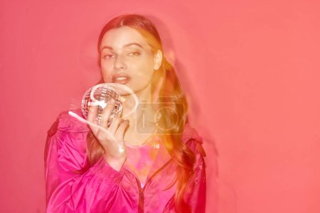Téléchargez les photos : A young woman in a pink dress elegantly holds a mirror ball, creating a dreamy and magical atmosphere in a studio with a pink background. - en image libre de droit