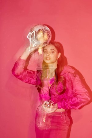 Téléchargez les photos : A stylish young woman in her 20s, wearing a pink shirt, holding a shiny silver ball against a pink background. - en image libre de droit