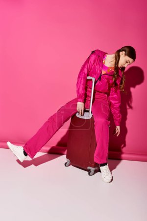 Una joven con estilo de unos 20 años se sienta encima de una maleta, encarnando la anticipación de la próxima aventura en un estudio con un fondo rosa.