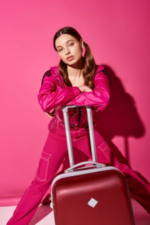A stylish woman in her 20s posing with a suitcase against a vibrant pink wall, exuding elegance and wanderlust vibes.