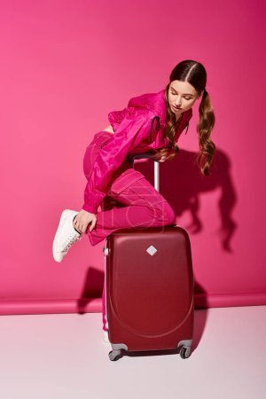A stylish young woman in her 20s sits on a red suitcase, wearing a fashionable suit in a studio with a pink background.