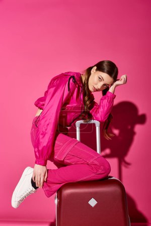 A stylish young woman in her 20s sitting on top of a red suitcase against a pink background.