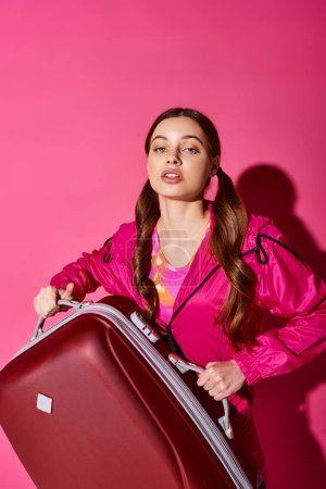 A stylish young woman in her 20s, wearing a pink jacket, holding a red suitcase against a pink backdrop.