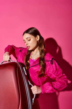 Téléchargez les photos : A stylish woman in her 20s clad in pink holds a suitcase in a studio with a pink backdrop, exuding a sense of adventure. - en image libre de droit