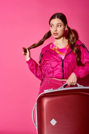 Photo for A young, stylish woman in her 20s wearing a pink jacket holding a red suitcase in a studio setting with a pink background. - Royalty Free Image
