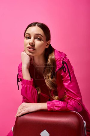 Una chica joven y elegante de unos veinte años se sienta encima de una maleta de color rojo brillante en un estudio, sobre un fondo rosa.