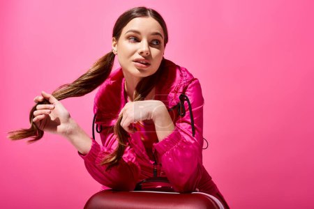 A stylish young woman in her 20s sitting atop a red suitcase against a pink studio backdrop.