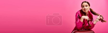 Photo for A young beautiful woman in her 20s sitting atop a red suitcase in a studio with a pink background. - Royalty Free Image