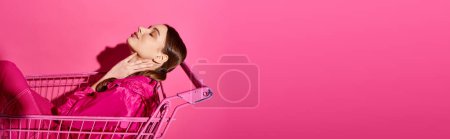 Una mujer con estilo en sus 20 años se sienta con los ojos cerrados en un carrito de compras sobre un fondo de estudio rosa.