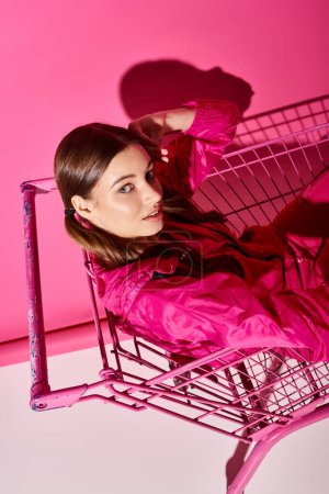 A young, stylish woman in her 20s lies gracefully inside a shopping cart in a vivid pink room, exuding a sense of dreamy euphoria.