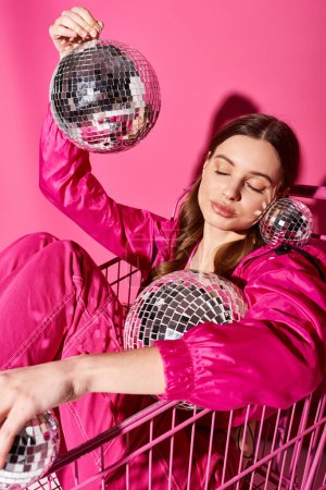 Foto de Una joven y hermosa mujer de unos 20 años, elegante y de moda, se sienta con confianza en un carrito de compras contra un vibrante telón de fondo rosa. - Imagen libre de derechos