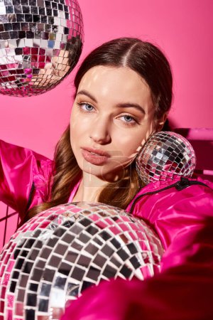 Foto de Stylish young woman in her 20s holding two disco balls, wearing a pink dress, set against a vibrant pink background. - Imagen libre de derechos