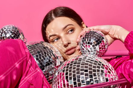 A stylish young woman in her 20s, dressed in a pink outfit, holds a reflective mirror ball in a vibrant studio setting.