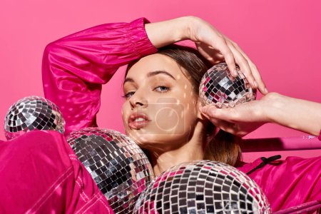 Téléchargez les photos : A stylish young woman in her 20s wearing a pink outfit, holding disco balls in a vibrant studio setting with pink background. - en image libre de droit
