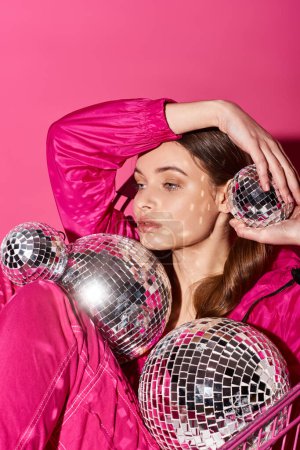 Foto de A stylish woman in her 20s wearing a pink outfit holds multiple shiny disco balls in a studio with a pink background. - Imagen libre de derechos