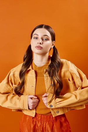 Eine stylische junge Frau in ihren Zwanzigern posiert in einem Atelier, trägt eine leuchtend gelbe Jacke und eine trendige orangefarbene Hose vor orangefarbenem Hintergrund..