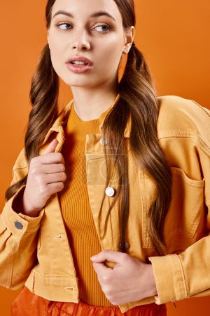 Foto de Una mujer joven y elegante de unos 20 años con el pelo largo, con una chaqueta amarilla, posando en un estudio sobre un fondo naranja. - Imagen libre de derechos