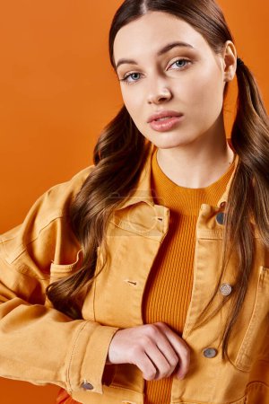 Foto de Una mujer con estilo de unos 20 años con el pelo largo posando en una chaqueta amarilla sobre un fondo naranja. - Imagen libre de derechos