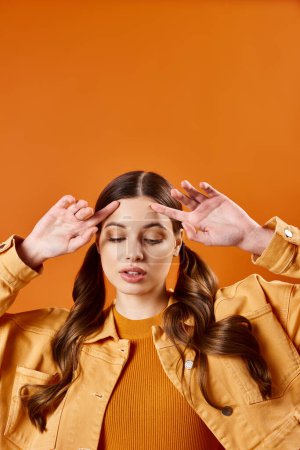 Foto de Una joven de unos 20 años con las manos en la cabeza en un estudio con un fondo naranja, que se ve abrumada. - Imagen libre de derechos