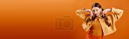Téléchargez les photos : Jeune femme dans la vingtaine portant une robe jaune vif avec ses mains sur la tête dans un cadre de studio sur un fond orange. - en image libre de droit