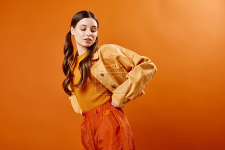 A stylish young woman in her 20s wearing a vibrant yellow shirt and pants poses in a studio with an orange background.