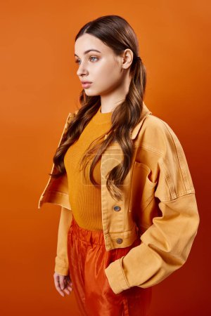 A stylish young woman in her 20s stands gracefully against a vibrant orange backdrop in a studio setting.