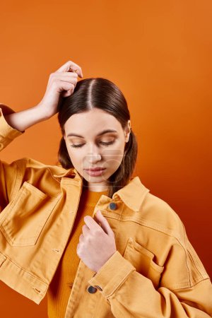 A stylish woman in her 20s, wearing a yellow jacket, elegantly holds her hair against an orange backdrop in a studio setting.