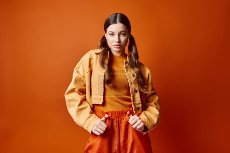 Foto de Una mujer joven y elegante de unos 20 años se para con confianza frente a un fondo naranja brillante en un entorno de estudio. - Imagen libre de derechos